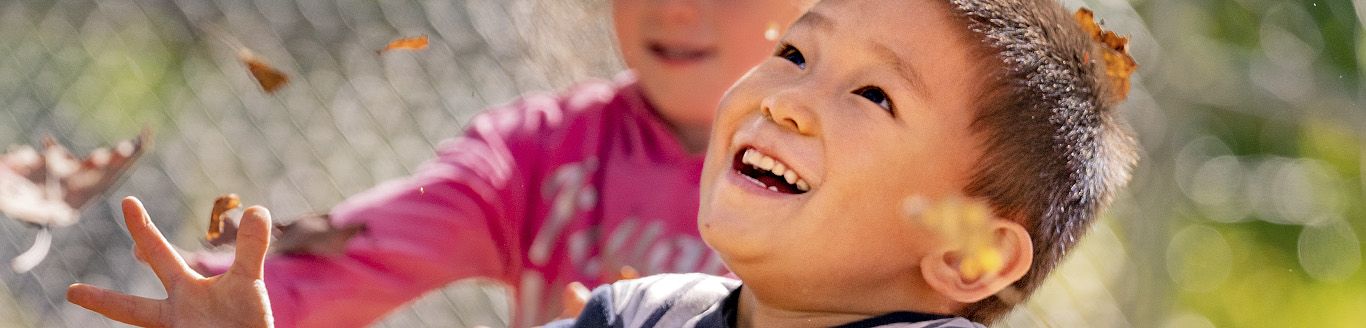 Two children playing outdoors, one laughing with leaves in their hair and hands.