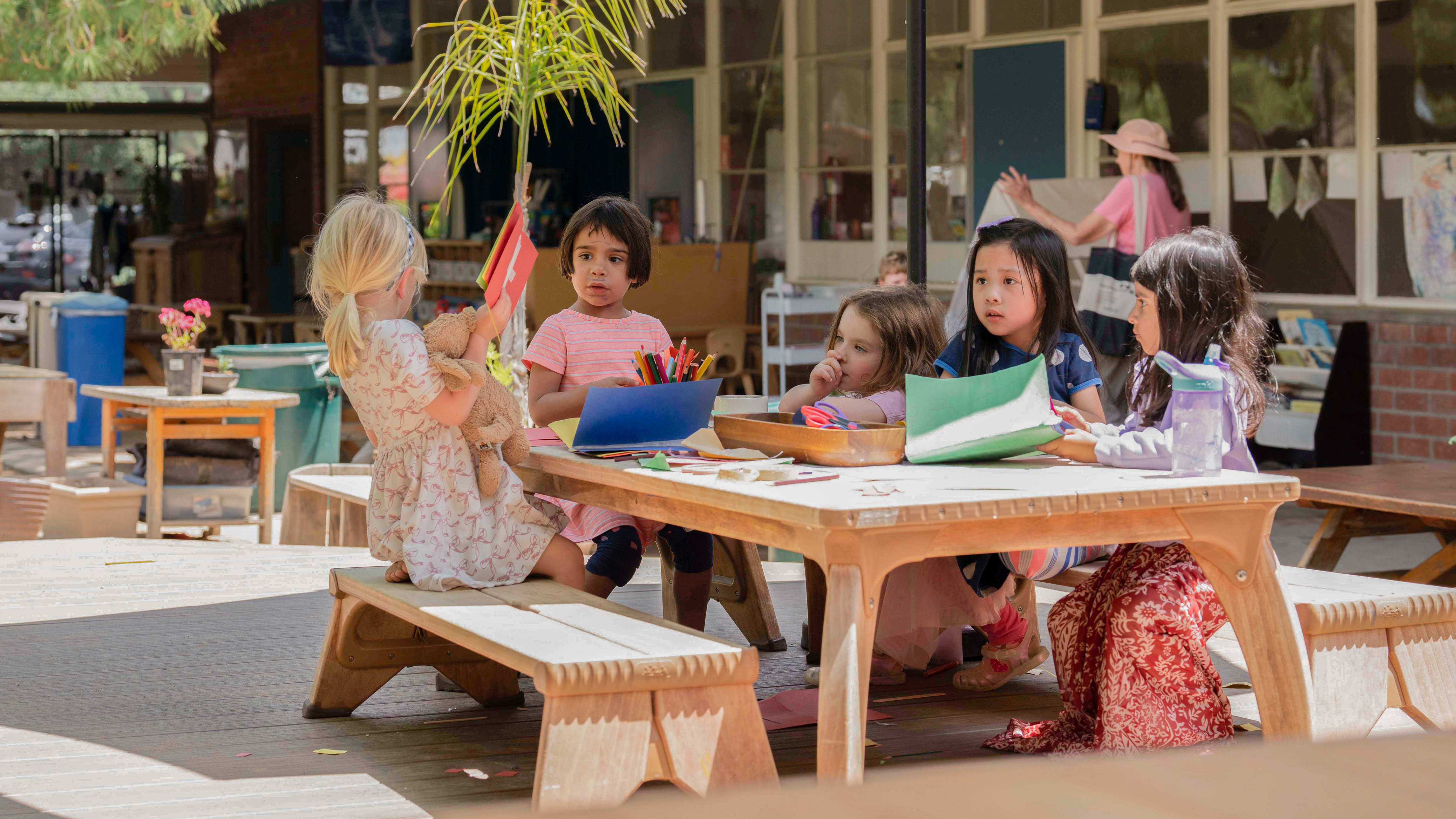 Five young children sit at a wooden picnic table outdoors, drawing and crafting.