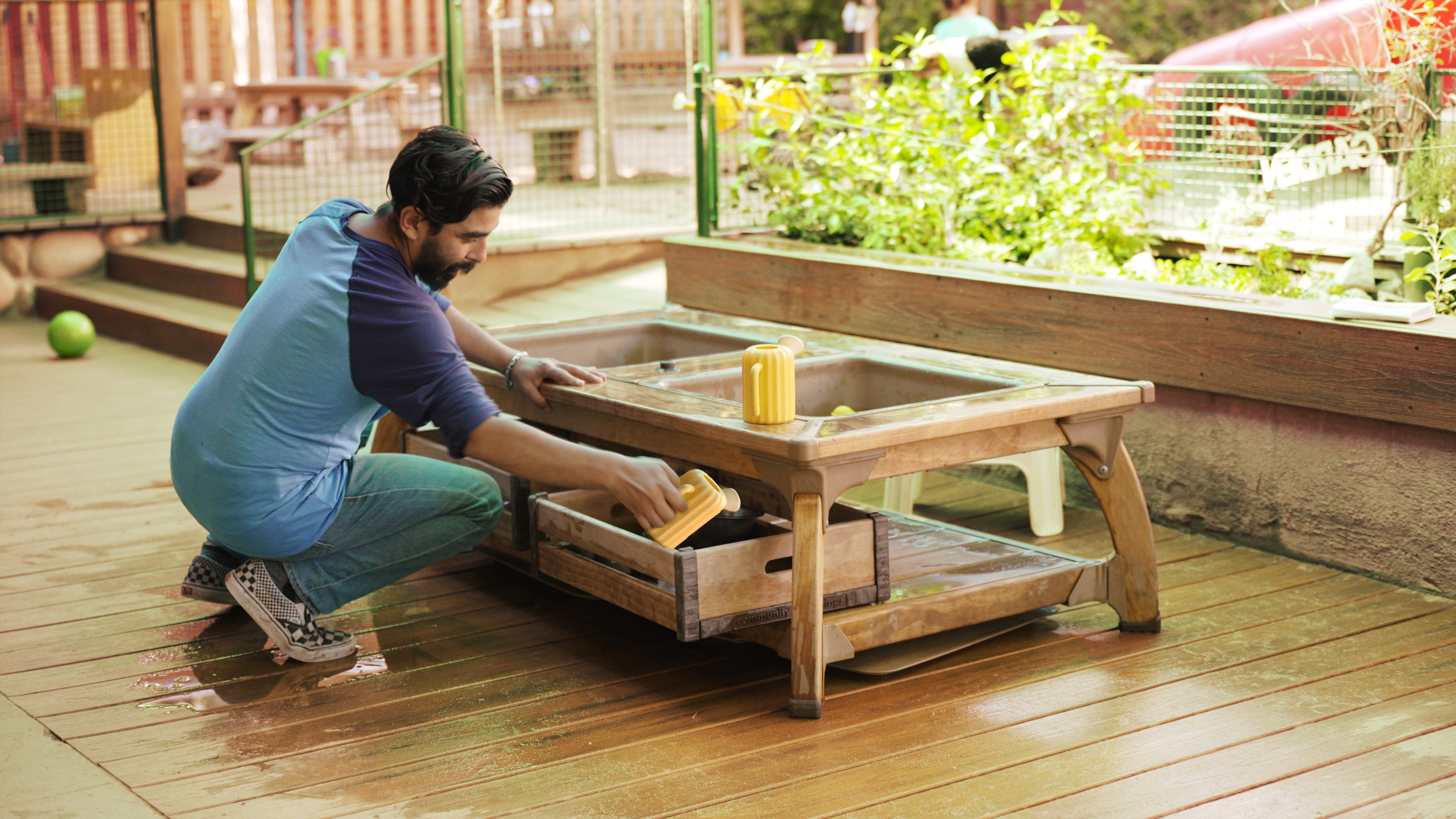Man crouching, placing a yellow toy into a wooden storage drawer under a water play table.