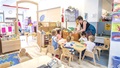 Teacher helping five young children seated at a table in a bright classroom with wooden furniture.