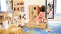 Toddler in a wooden wagon looking at her reflection in a mirror, with a teacher in the background.