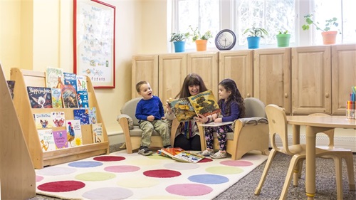 Teacher reads to two children sitting in gray upholstered chairs on a colorful polka-dot rug.