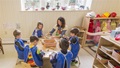 Teacher and five preschoolers in blue smocks making clay creations at a round wooden table.