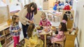 Teacher helping four young girls in dress-up clothes play with toy food at a round table.