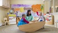 Teacher and two young children playing with a wooden rocking boat in a classroom.