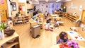 Children and a teacher in a busy Montessori classroom with wooden furniture and learning materials.