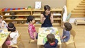A teacher sits with four children at a table, talking to them during lunch time.