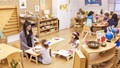A teacher and children working at tables and on the floor in a bright Montessori classroom.