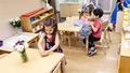 Children seated at light wood tables in a classroom, engaged in various activities.
