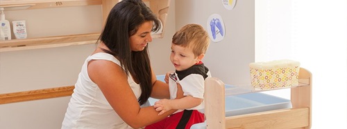 Teacher helping a young boy in a white and black shirt with red pants at a changing table.