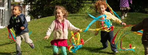 Three children running on a grassy hill, holding colorful ribbons that stream behind them.