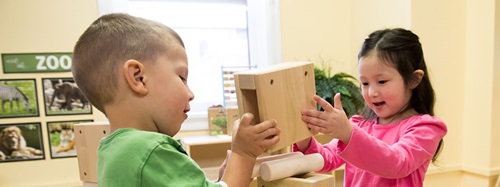 Two young children playing together with wooden building blocks in a classroom.