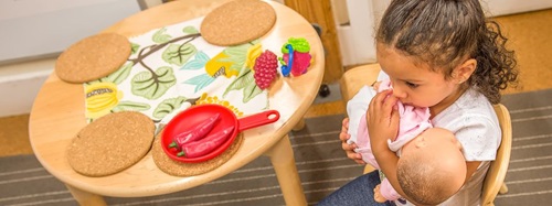 A young girl in a white shirt holds a baby doll while sitting at a small wooden table.