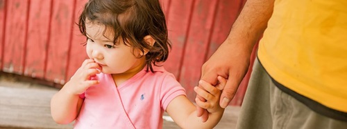 A young child in a pink shirt holds an adult's hand, with a red wall in the background.