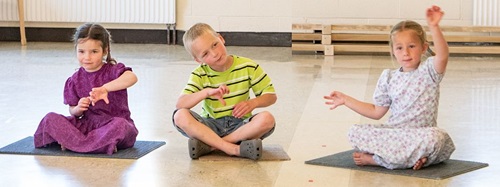 Three children sitting on mats on the floor, gesturing with their hands.