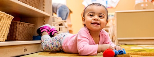 Happy infant in a pink shirt crawling on a rug, looking at the camera.