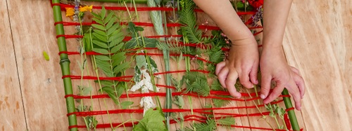 Child's hands weaving natural elements like leaves and flowers into a bamboo frame with red yarn.