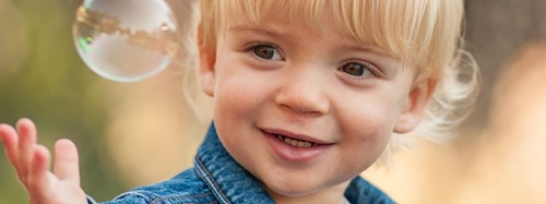 A smiling toddler with blonde hair and a denim jacket reaches for a bubble.