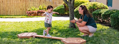 A toddler balances on a low wooden beam while a woman crouches nearby, smiling and ready to assist.