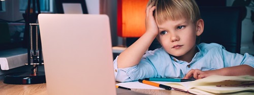 A young boy with blonde hair rests his head on his hand, looking bored at a laptop screen.
