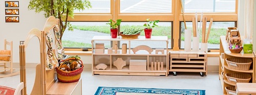 A bright classroom with wooden shelves, blocks, and baskets of toys by large windows.