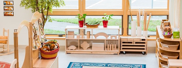 A bright classroom with wooden shelves, blocks, and baskets of toys by large windows.