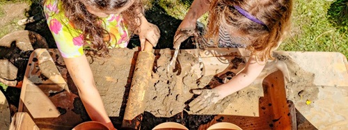 Two young girls playing on a mud kitchen table, one is using a rolling pin and the other one is using a fork.