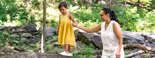 A woman holds a young girl's hand as the girl balances on a fallen log in a wooded area.