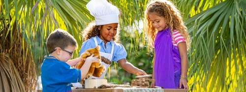 Three children playing outdoors, one in a chef's hat and apron, with a teddy bear and wood pieces.
