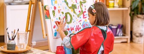 A young girl in a red smock finger painting on an easel.