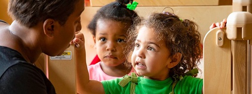 Two young children and an adult interacting in a classroom setting, with one child looking up intently.