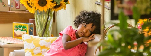 A young girl with curly hair rests her head on a chair, looking at the camera. Sunflowers are in the background.