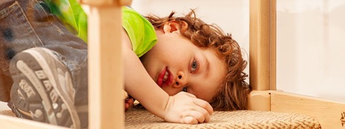 A young child with curly hair lies on a carpeted surface, looking up at the camera.