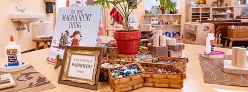 A classroom table with craft supplies, including glue, wooden blocks, marbles, and a book titled "The Most Magnificent Thing."