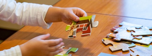 Child's hands holding a puzzle piece with a red house and green roof, other pieces on a wooden table.