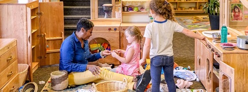 Teacher and two children playing in a classroom with wooden furniture and play kitchen.