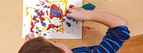 Child squeezing red, yellow, and blue paint from bottles onto white paper.
