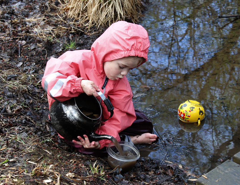 Child playing with old teapot  outdoors by a large puddle of water