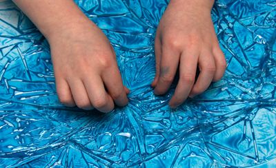 Hands pulling at Saran wrap over a sheet of paper with wet tempera paint