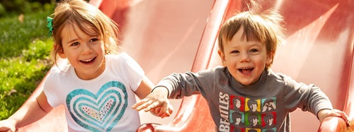 Two happy children, a girl and a boy, smiling while playing on a red slide outdoors.