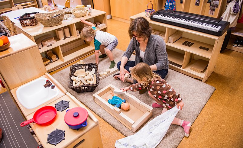 Children creating doll be with unit blocks while teacher sits on the floor observing