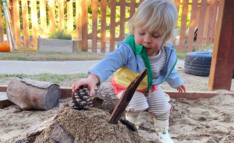 Child adding a pinecone to the top of a sand castle in sandbox outdoors