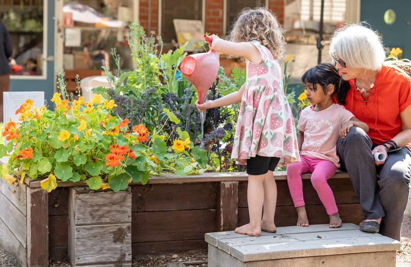 A child watering plants with a teacher and child watching