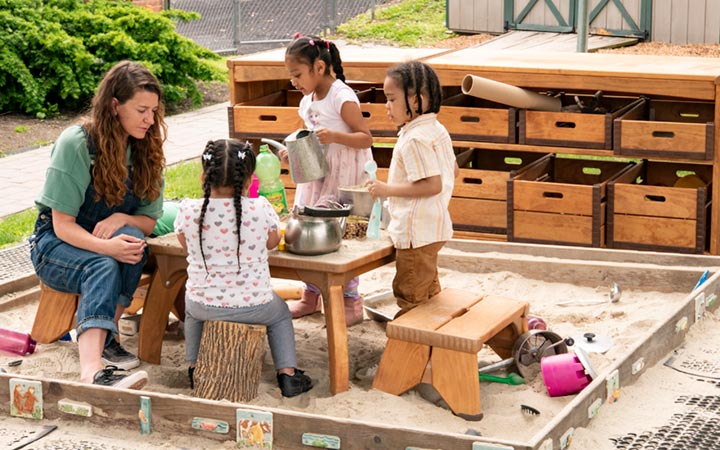Teacher seated at Outlast Play Table in sandbox watching the children making sand cakes
