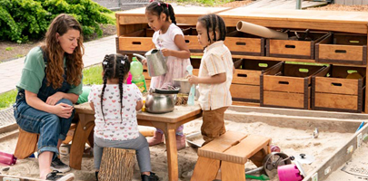 Teacher seated at Outlast Play Table in sandbox watching the children making sand cakes