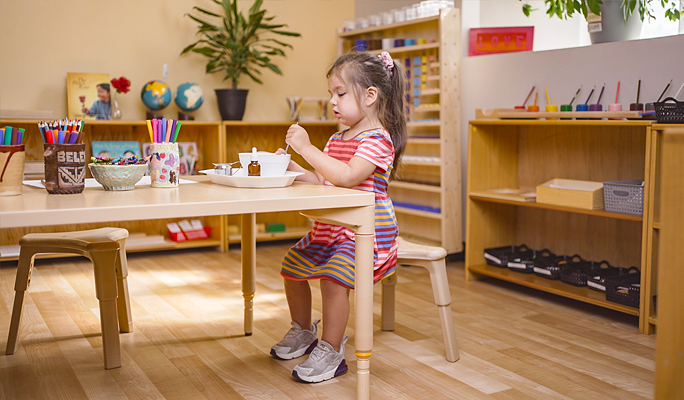 Young child in a striped dress sitting at a light wood table, engaged in an activity with small bowls and a dropper.