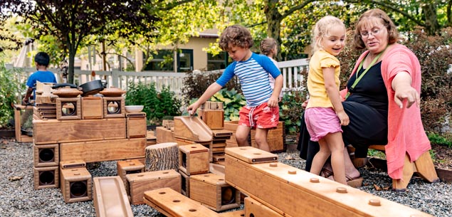 Children and an adult playing with large wooden outdoor blocks in a gravel play area.