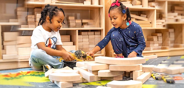 Two children playing with wooden blocks and toy animals on a colorful rug.