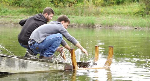 Two men on a wooden raft testing a wooden structure in a pond, splashing water.