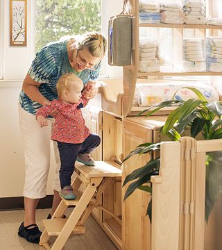 A teacher is helping a toddler up the steps to the changing table in the sunlit changing area of a child care setting.
